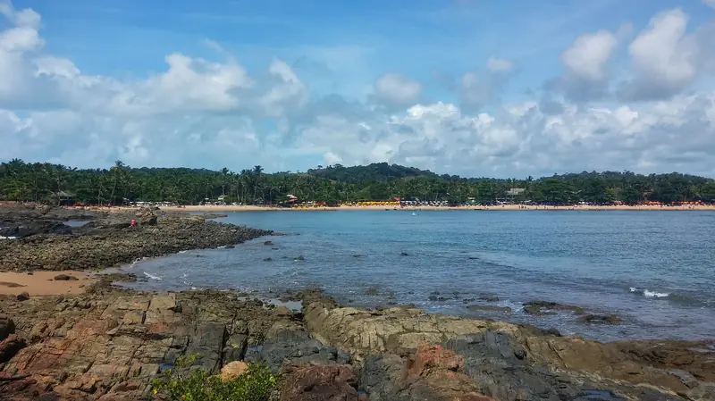 Mar azul calmo numa enseada com pedras em primeiro plano e a faixa de areia da praia ao fundo, com guarda-sóis e coqueiros