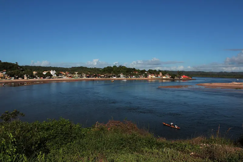 Rio largo e azul com uma canoa no meio e, na outra margem, a fileira de casas coloridas da cidade junto à mata