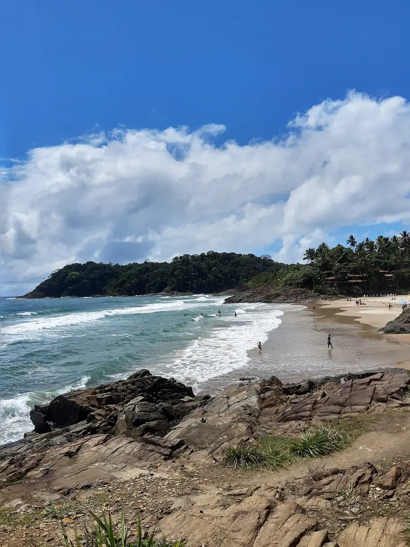 Ondas quebrando numa praia de areia clara entre pedras e um morro de mata, com surfistas na água sob céu azul