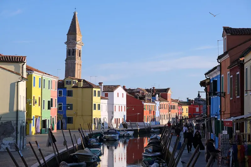 Canal de Burano com casas pintadas de amarelo, azul e vermelho, barcos atracados e o campanário inclinado de San Martino ao fundo