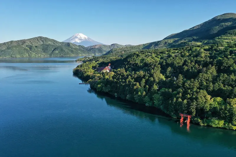 Lago Ashi visto do alto, com floresta na margem, o portal vermelho do santuário de Hakone na água e o monte Fuji nevado ao fundo