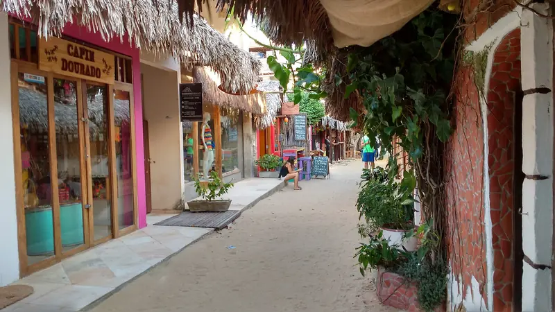 Rua de areia da vila com lojas coloridas e beirais de palha, entre plantas e muros de tijolo