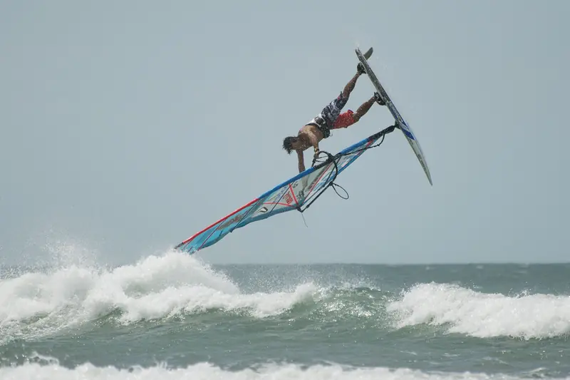 Windsurfista no ar, de cabeça para baixo sobre a prancha e a vela, acima de uma onda quebrando