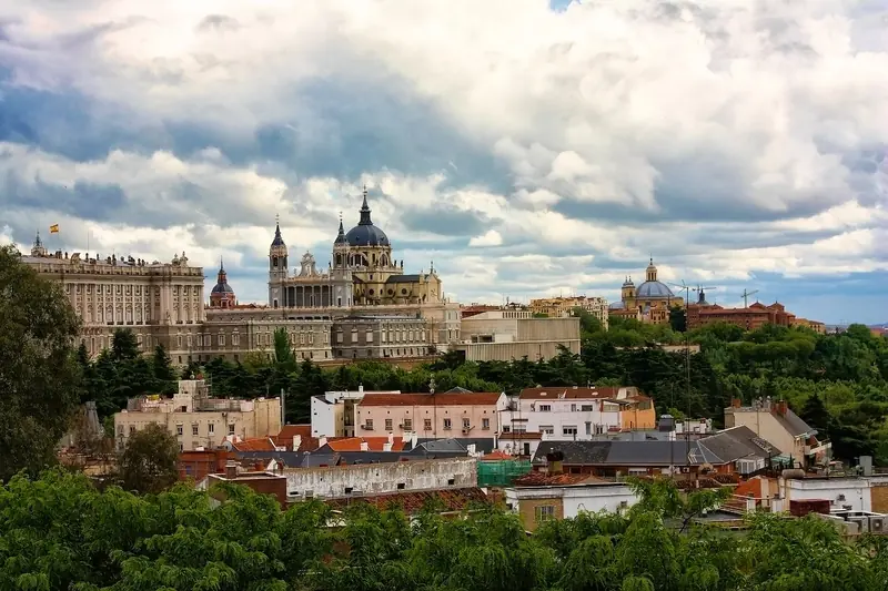 Palácio Real e catedral de Almudena vistos do parque do Templo de Debod, acima dos telhados e das árvores, sob céu com nuvens