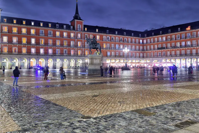 Plaza Mayor de Madri à noite, com o chão molhado refletindo as arcadas iluminadas e a estátua equestre de Felipe III