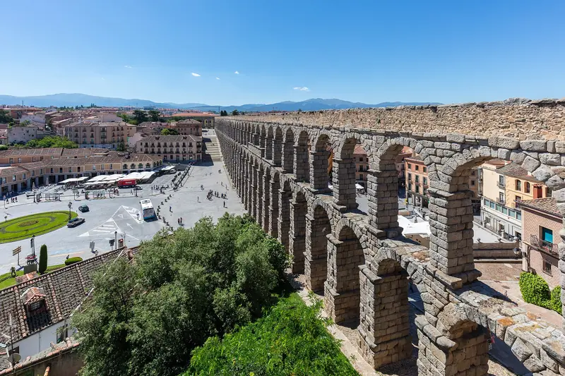 Arcos de pedra do aqueduto romano de Segóvia vistos do alto, com a praça do Azoguejo e as montanhas ao fundo