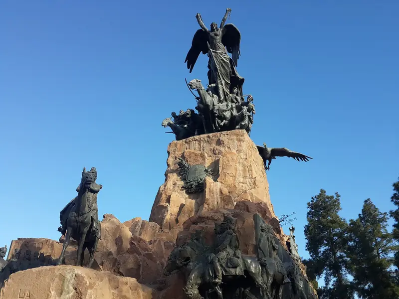 Monumento de bronze sobre um bloco de pedra, com uma figura alada no topo e cavaleiros em volta, contra o céu azul