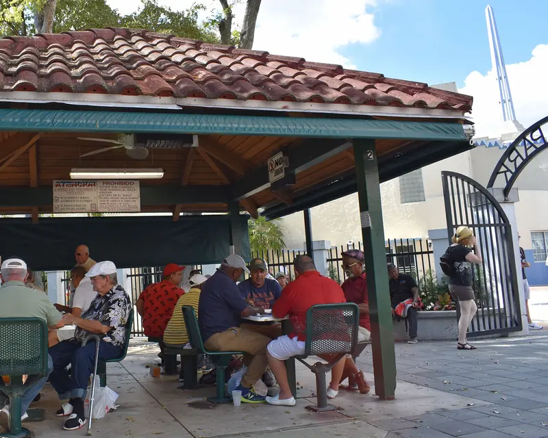 Homens jogando dominó sob uma cobertura de telhas de barro no Domino Park, com portão de ferro e prédios ao fundo
