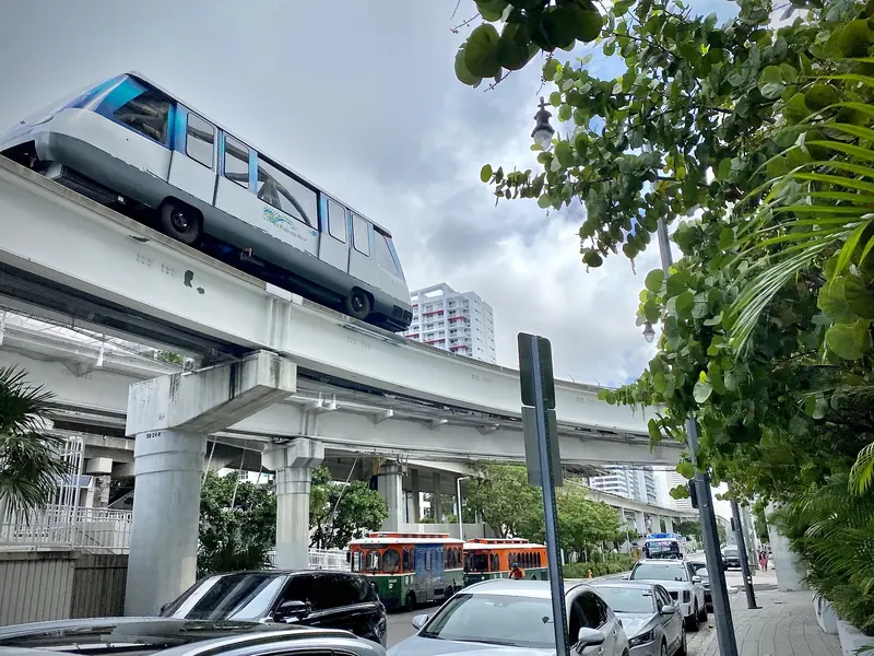 Vagão branco e azul do Metromover passando sobre o trilho elevado em Brickell, com trolleys estacionados embaixo e prédios ao fundo