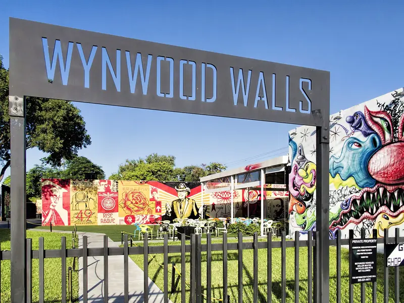 Portão de ferro com o letreiro Wynwood Walls, gramado e murais coloridos ao fundo, sob céu azul
