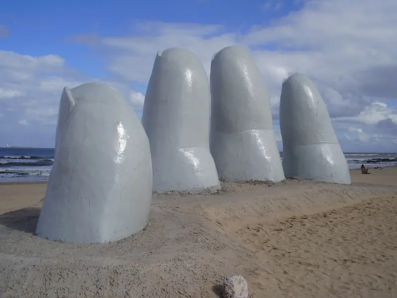 Os dedos gigantes da escultura La Mano saindo da areia da Playa Brava, com o mar e nuvens ao fundo