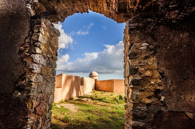 Muralha de pedra com uma abertura em arco que enquadra a guarita e o muro claro da fortaleza sob céu azul