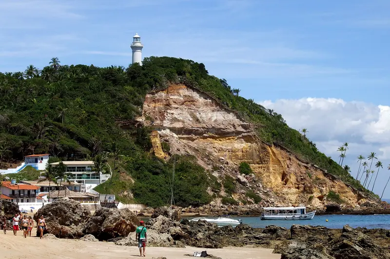 Morro coberto de mata com uma encosta de barro avermelhado e o farol branco no alto, sobre pedras, casas e um barco na água