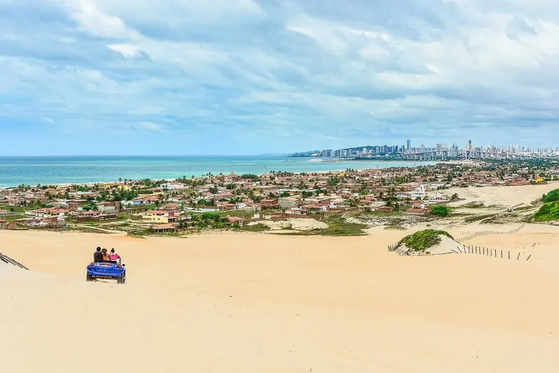 Buggy azul descendo uma duna de areia clara em Genipabu, com casas, o mar e os prédios de Natal ao fundo