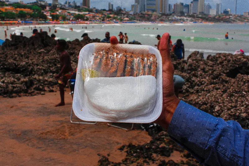 Mão segurando uma bandeja com tapioca branca e espetinhos de peixe frito, na praia, com banhistas e pedras ao fundo