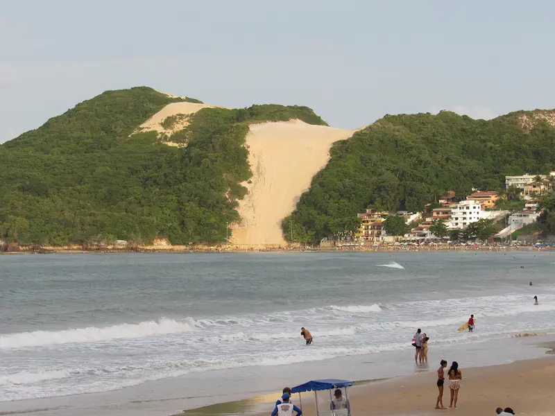 Duna alta de areia clara entre dois morros cobertos de mata, sobre a praia, com banhistas no mar em primeiro plano