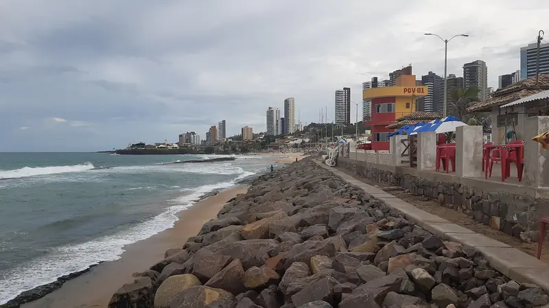 Calçadão sobre um enrocamento de pedras junto a uma faixa de areia estreita, com o mar à esquerda e prédios altos ao fundo, em dia nublado