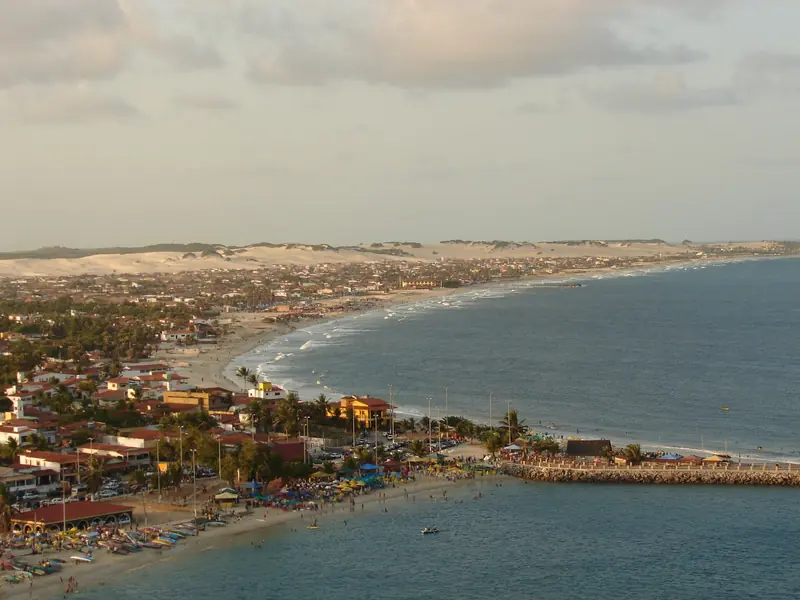Vista do alto de uma praia urbana curva, com barcos na areia e um molhe, e uma faixa de dunas claras no horizonte