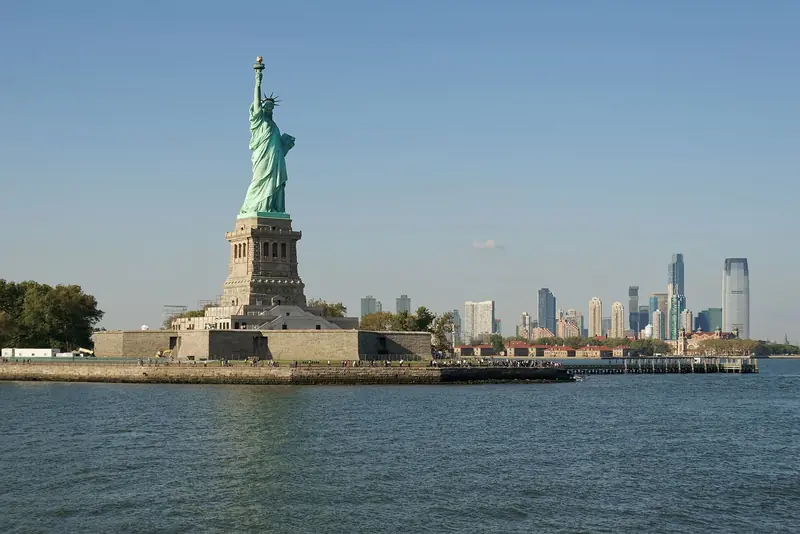 Estátua da Liberdade sobre o pedestal na Ilha da Liberdade, vista da água, com os prédios de Jersey City à direita