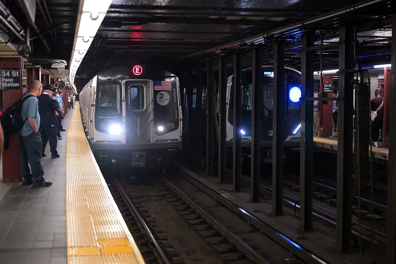Trem da linha E chegando à plataforma da estação 34 St-Penn Station, com passageiros esperando à esquerda