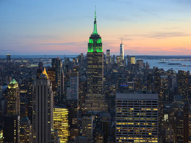 Empire State iluminado de verde ao entardecer visto do alto, com o One World Trade Center ao fundo