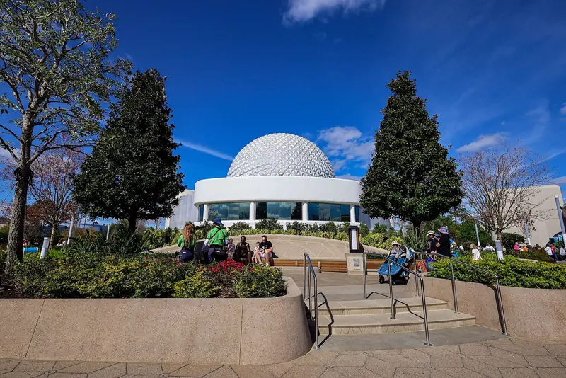 Esfera geodésica da Spaceship Earth atrás de um prédio branco redondo, vista de um jardim com escadaria e duas árvores altas