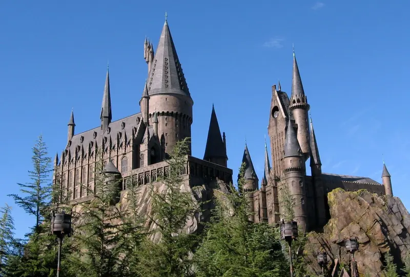 Castelo de Hogwarts sobre rochas, com torres pontiagudas e pinheiros em primeiro plano, sob céu azul