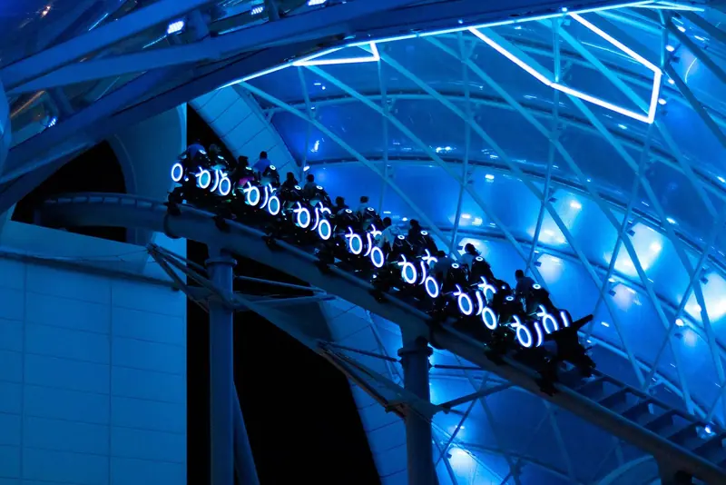 Trem de motos iluminadas em azul da Tron Lightcycle / Run passando sob a cobertura de vidro, à noite