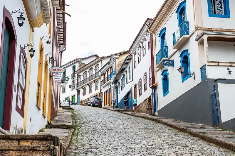 Rua de pedra inclinada com casarões coloniais de janelas azuis e amarelas