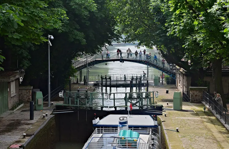 Eclusa do Canal Saint-Martin com barco esperando, passarela de ferro ao fundo e árvores dos dois lados