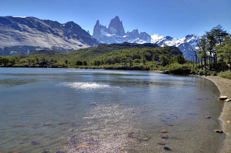 Lago de água clara com bosque em volta e o Fitz Roy nevado ao fundo, sob céu limpo