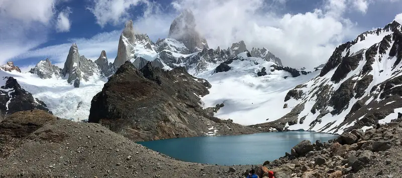 Laguna de los Tres azul-turquesa ao pé do Fitz Roy e de agulhas de granito cobertas de neve, com caminhantes nas pedras em primeiro plano