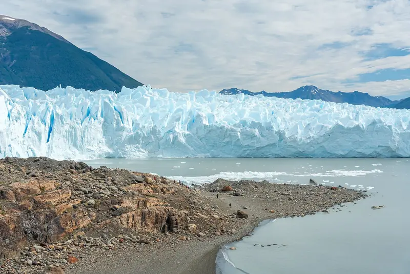 Parede do Perito Moreno ao fundo e praia de cascalho com caminhantes pequenos em primeiro plano