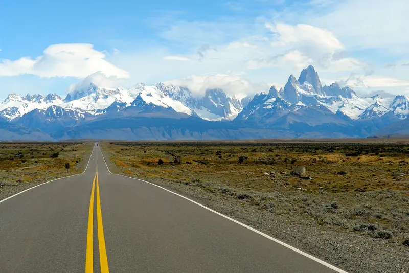 Estrada asfaltada reta com faixa amarela dupla atravessando a estepe, com o maciço do Fitz Roy nevado ao fundo