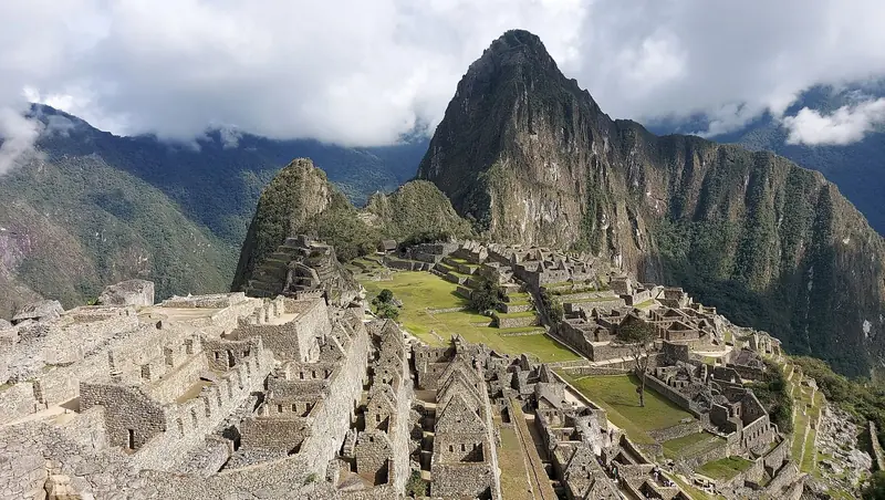 Ruínas de Machu Picchu vistas do alto, com terraços, casas de pedra e a praça central gramada, e o pico de Huayna Picchu ao fundo sob nuvens