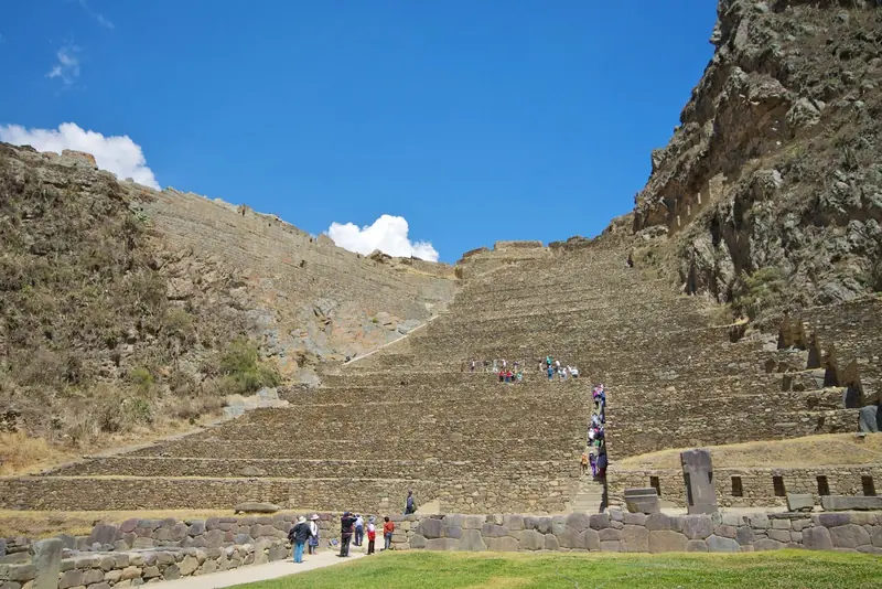 Terraços de pedra da fortaleza de Ollantaytambo subindo a encosta, com visitantes nas escadarias e muros incas na base