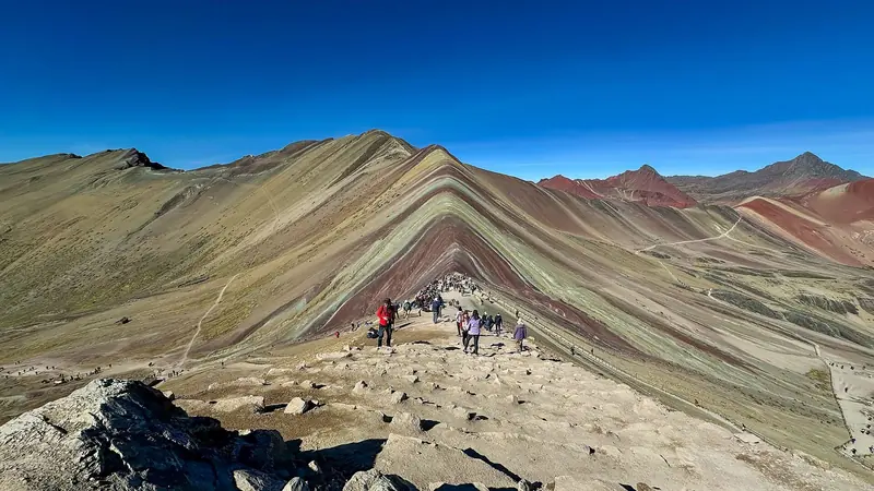 Faixas de cor vermelha, amarela e verde nas encostas da Vinicunca, com uma fila de visitantes na trilha do mirante sob céu azul