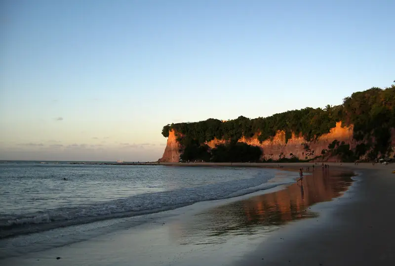 Faixa de areia molhada refletindo uma falésia alaranjada coberta de mata, com o mar calmo à esquerda, no fim da tarde