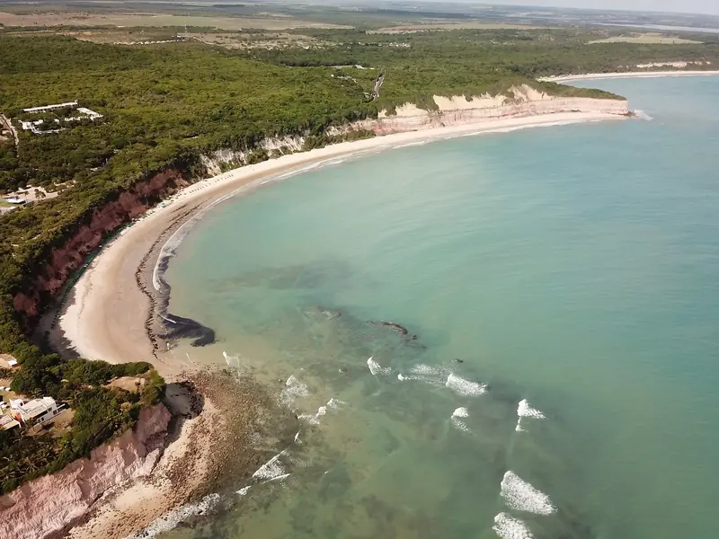 Vista aérea da Baía dos Golfinhos, em Pipa: uma enseada curva de areia clara ao pé de falésias cobertas de mata, com o mar verde-claro