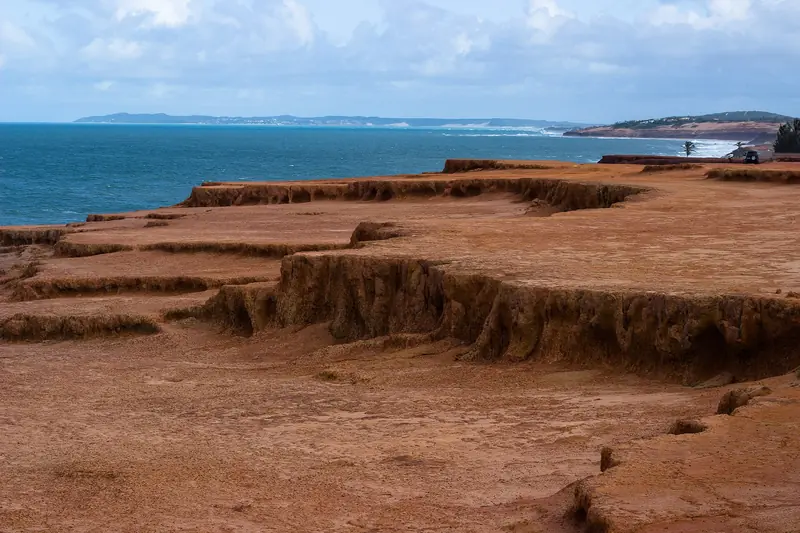 Platô de terra vermelha com degraus de erosão em primeiro plano e o mar azul ao fundo, com uma ponta de terra verde à direita