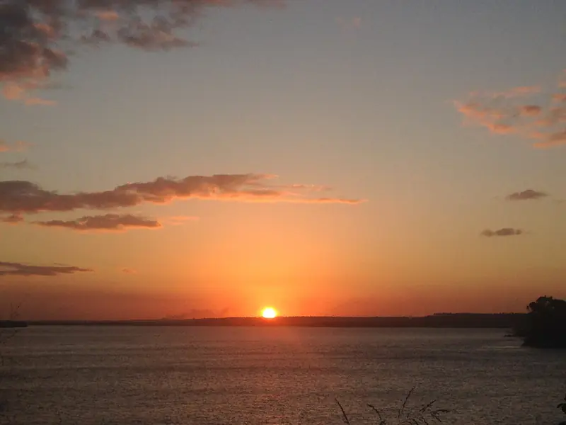 O sol baixo tocando o horizonte sobre a água larga e calma da lagoa, com o céu alaranjado e algumas nuvens