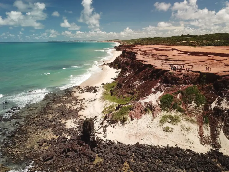 Vista aérea de uma falésia de terra vermelha com pessoas no alto, sobre uma praia de areia clara e pedras escuras, com o mar verde à esquerda