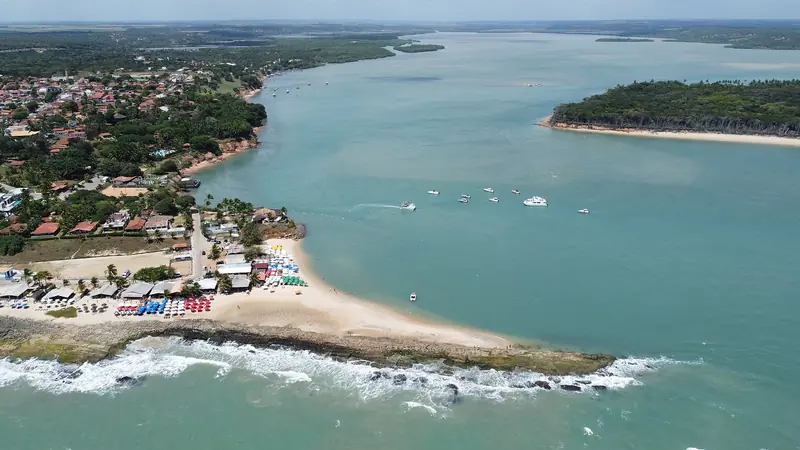 Vista aérea de uma vila com casas entre a vegetação e uma ponta de areia com barracas coloridas, onde a lagoa larga e verde-azulada encontra o mar