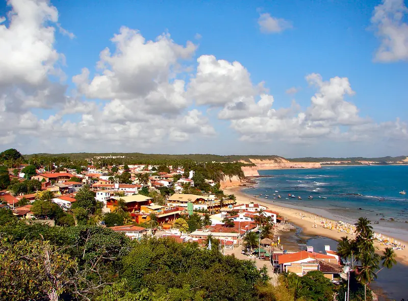 Vista do alto de uma vila de telhados vermelhos e muita vegetação, descendo até uma praia com guarda-sóis e barcos no mar azul