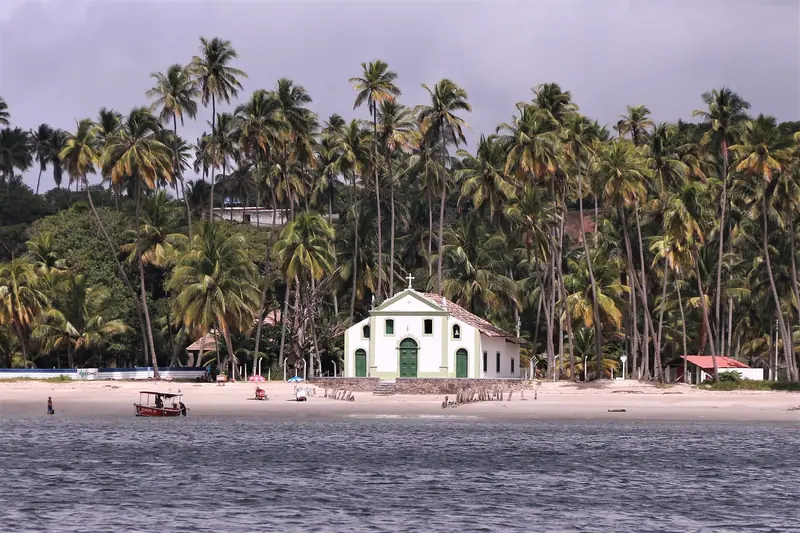 Capela branca de detalhes verdes na beira da areia, cercada de coqueiros altos, vista do rio com um barco vermelho ancorado