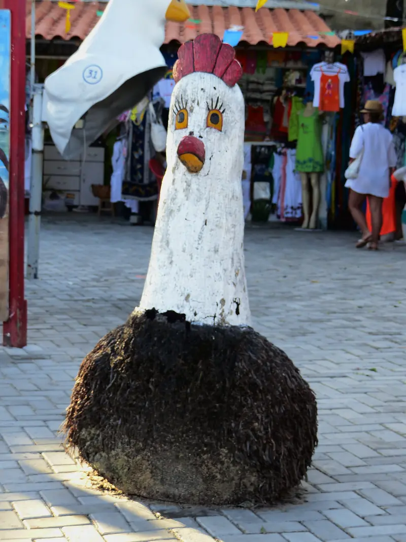 Escultura de galinha branca feita de tronco de coqueiro, com crista vermelha e olhos pintados, numa calçada de pedra diante de lojas de roupa