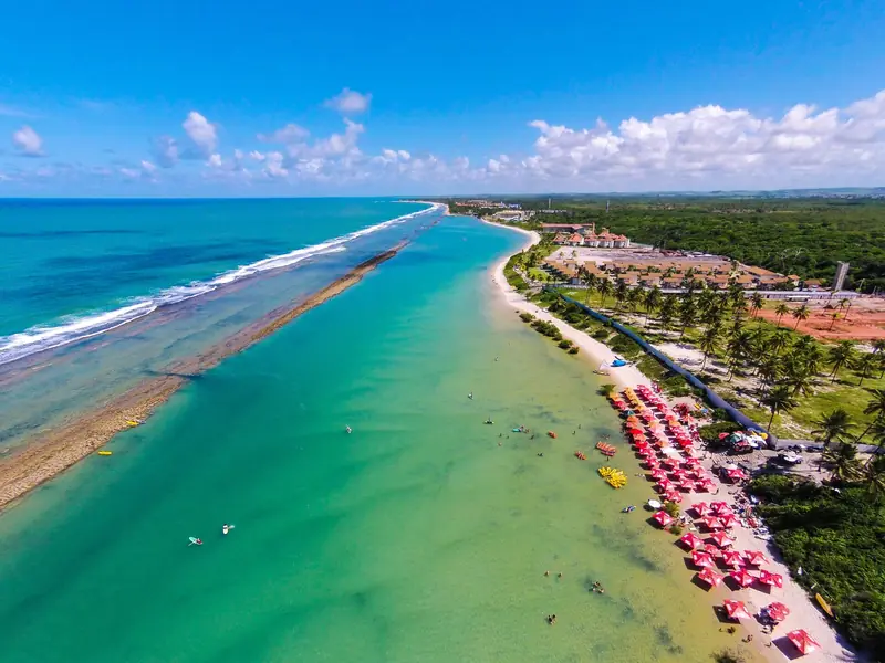 Vista aérea de Muro Alto, com o recife comprido separando o mar aberto, à esquerda, da água verde e calma junto à areia cheia de guarda-sóis vermelhos