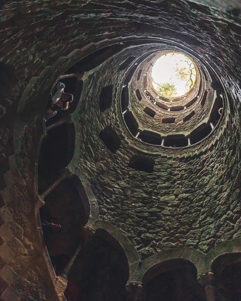 Vista de baixo do Poço Iniciático da Quinta da Regaleira, uma escada em espiral de pedra com a luz entrando pelo topo