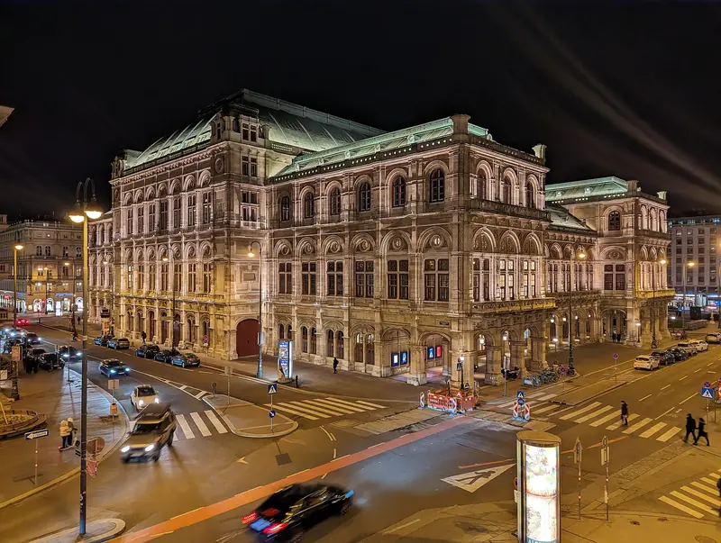 Fachada iluminada da Ópera Estatal de Viena à noite, vista do alto de uma esquina do Ring com carros passando