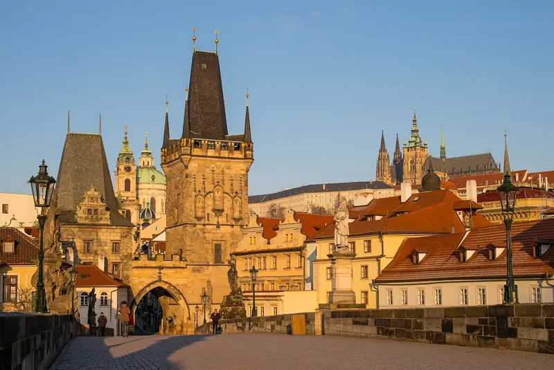Ponte Carlos quase vazia ao amanhecer, com a torre de Malá Strana e a catedral de São Vito ao fundo sob céu azul
